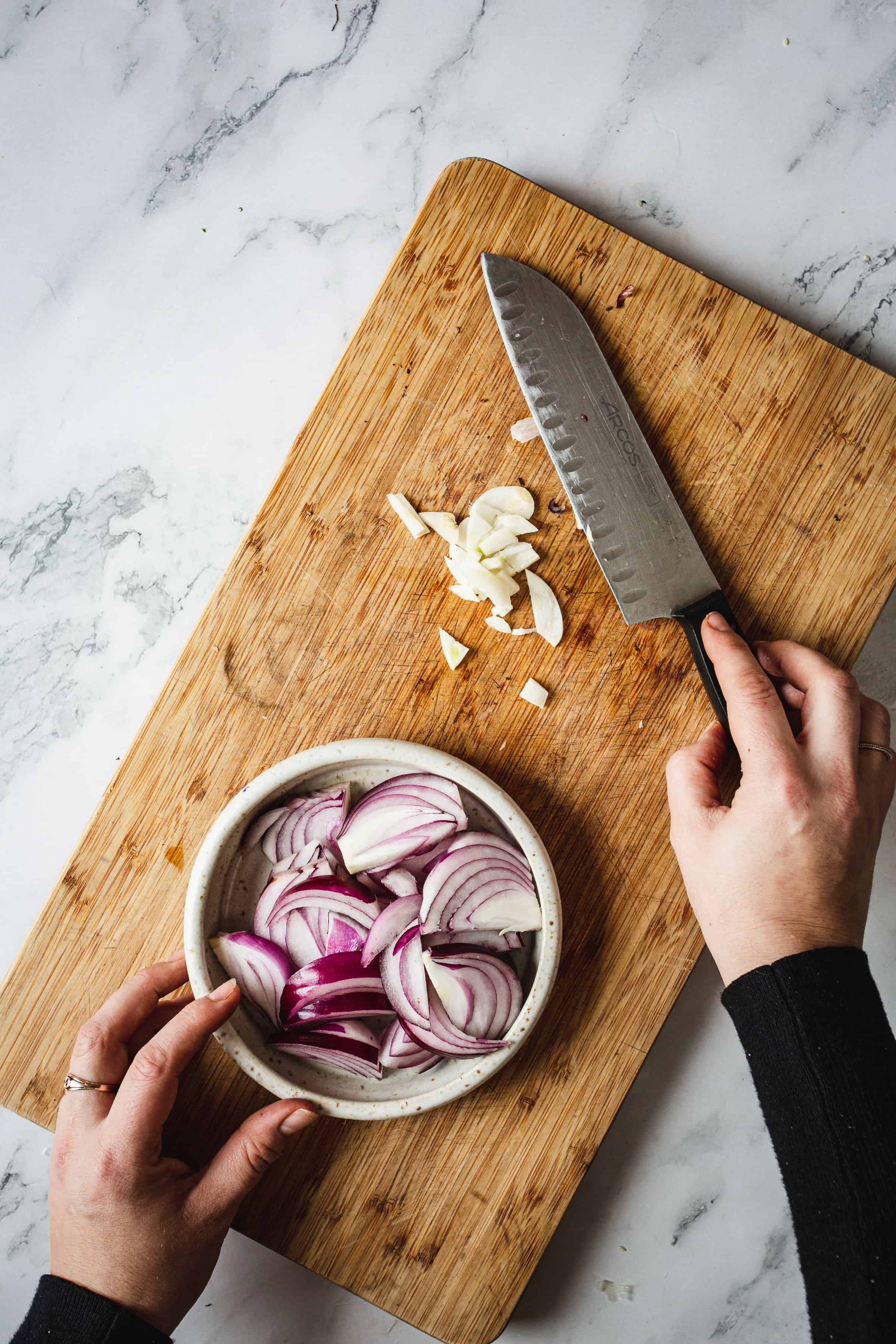 A person's hands arranging sliced red onions into a small bowl next to garlic pieces on a wooden cutting board with a chef's knife, on a marble countertop, near bell pepper bisque.