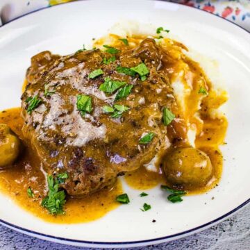 A plate of classic Salisbury steak topped with gravy and garnished with chopped parsley, accompanied by mashed potatoes and whole mushrooms, presented on a white plate with a blue rim.