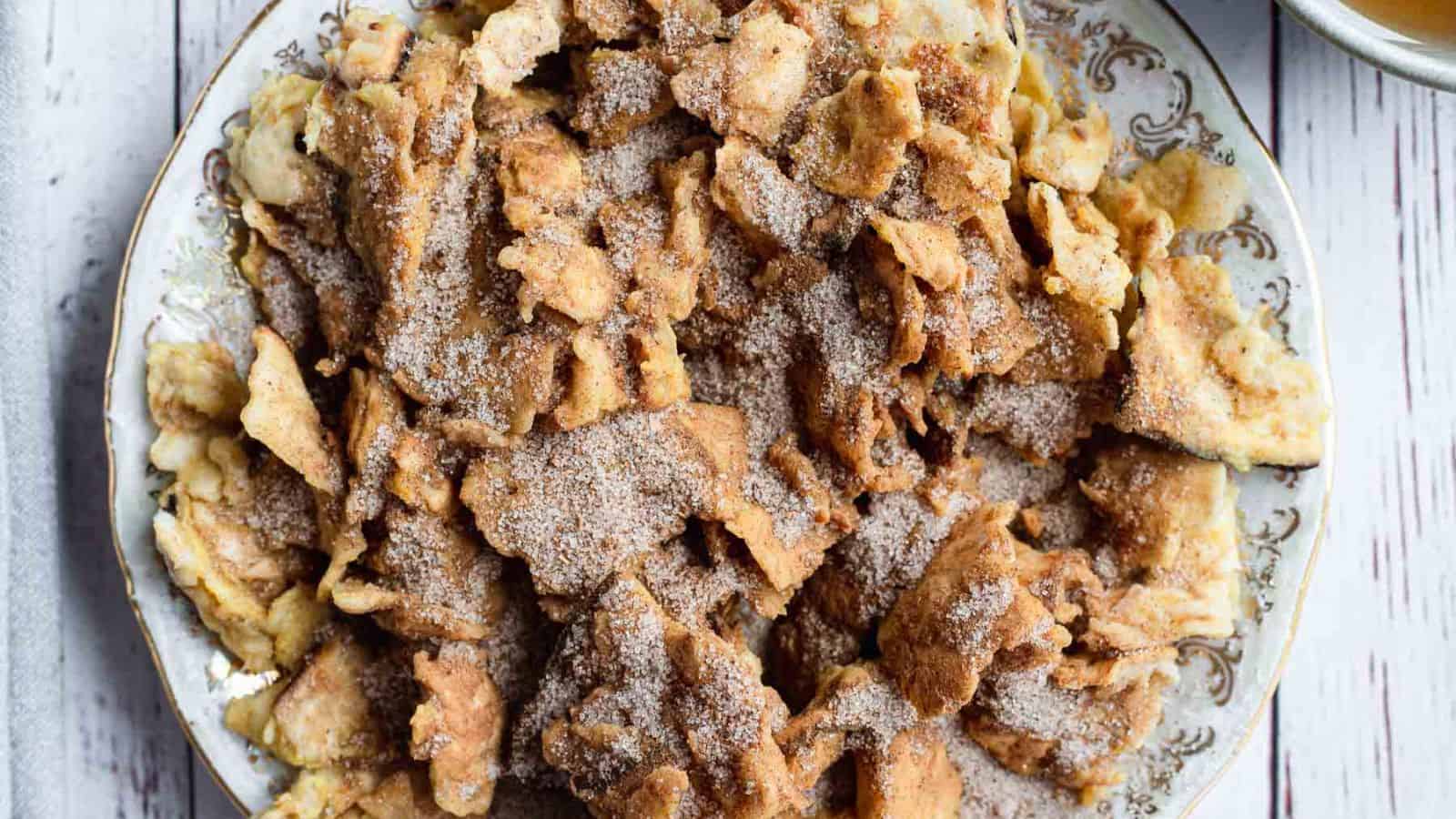 A plate filled with pieces of a dessert resembling Kaiserschmarrn, an Austrian shredded pancake dish. The pieces are golden-brown and dusted with powdered sugar. The plate has a decorative border and is placed on a white wooden surface, reminiscent of Grandma Recipes for Classic Passover dishes.