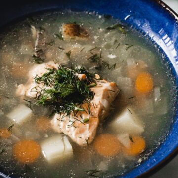 A bowl of clear broth soup with a piece of salmon, chopped carrots, potatoes, and garnished with dill, served in a blue rimmed bowl - straight from Grandma's secret recipes.