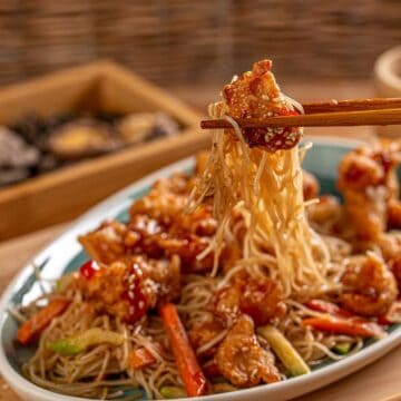 A plate of noodles with vegetables and pieces of fried chicken garnished with sesame seeds is being lifted with chopsticks. In the background, there is a wooden tray with mushrooms and a small cup, all set on a wooden surface, highlighting the harmonious relationship between flavors.