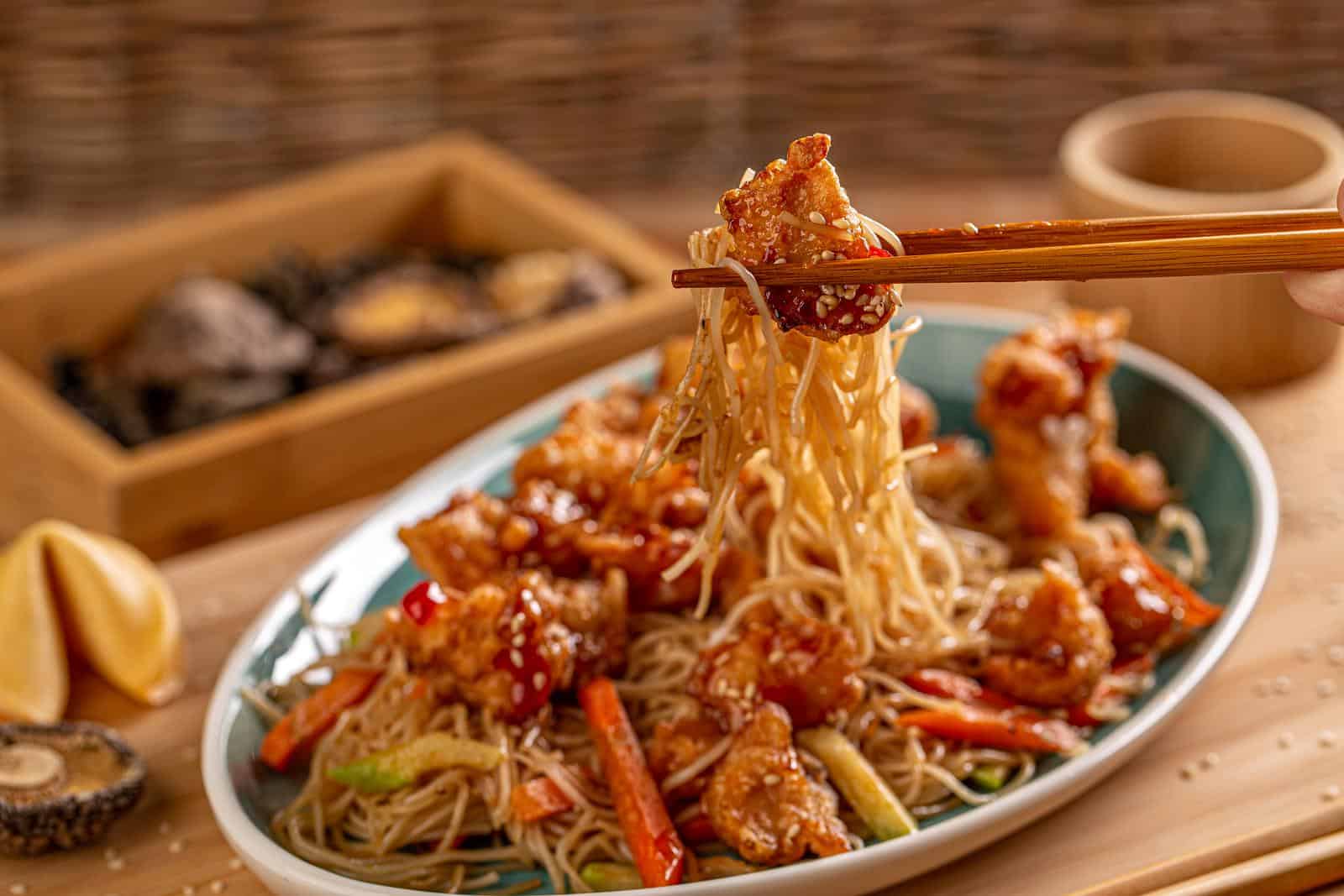 A plate of noodles with vegetables and pieces of fried chicken garnished with sesame seeds is being lifted with chopsticks. In the background, there is a wooden tray with mushrooms and a small cup, all set on a wooden surface, highlighting the harmonious relationship between flavors.