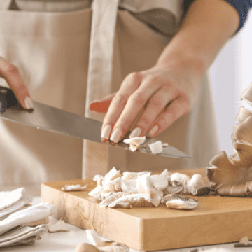 A person wearing an apron is slicing mushrooms on a wooden cutting board with a large kitchen knife. There are more mushrooms and a napkin on the table.