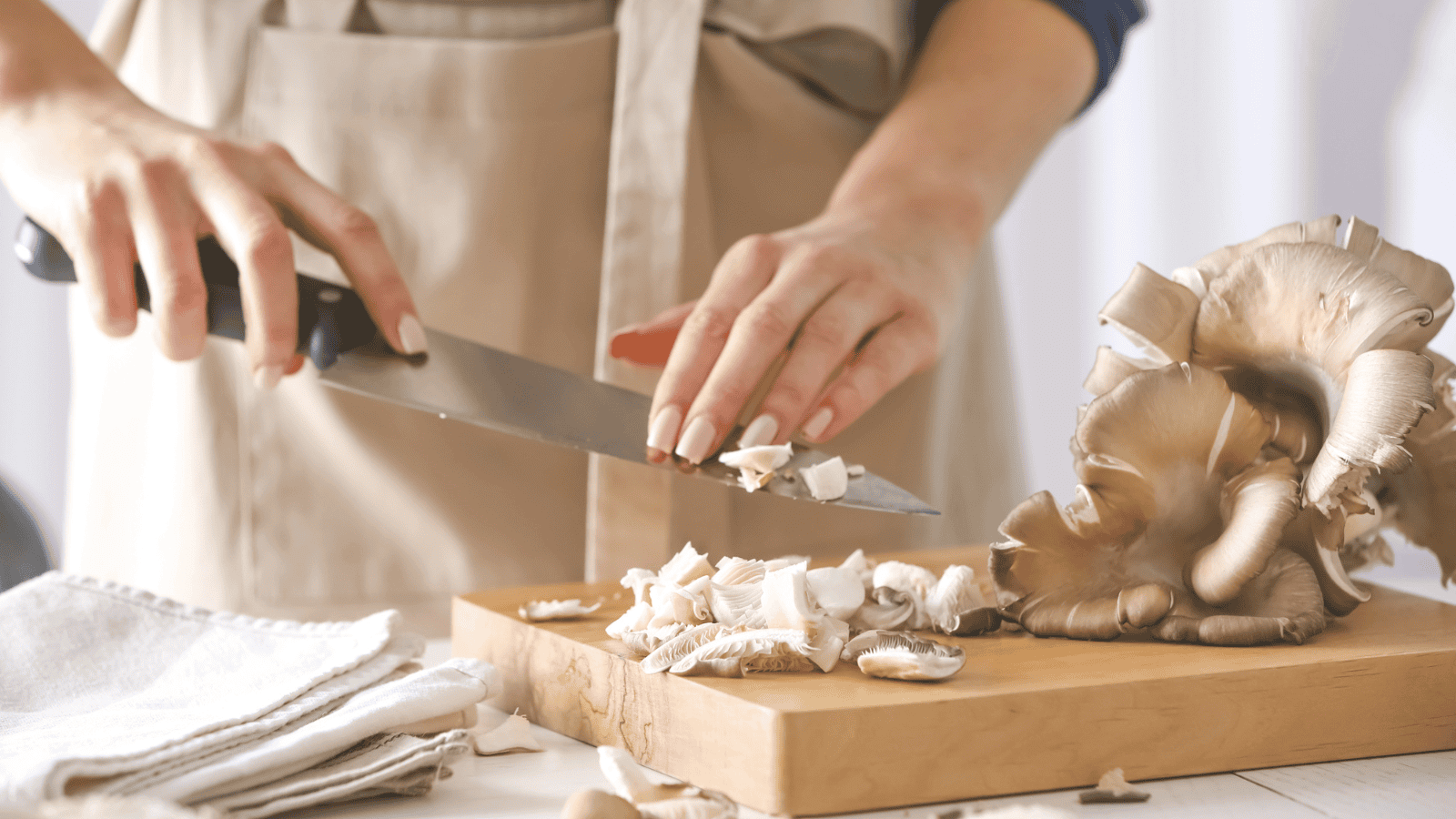 A person wearing an apron is slicing mushrooms on a wooden cutting board with a large kitchen knife. There are more mushrooms and a napkin on the table.