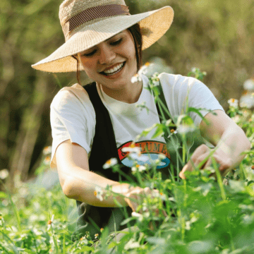 A person wearing a wide-brimmed hat and a white T-shirt is kneeling in a field of wildflowers, picking flowers and smiling. The background is blurred, showing more greenery and plants. The scene is sunny and suggests a calm, outdoor activity.