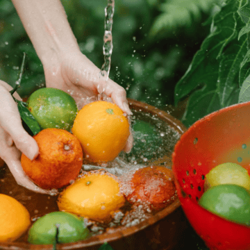 Two hands are seen washing various fruits, including oranges and limes, in a basin of water. Another red colander containing more limes is next to the basin. Green leafy foliage is visible in the background, dispelling any common mistakes about fruit cleanliness and preparation.
