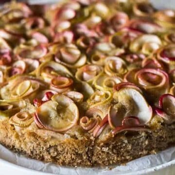 A close-up of a round apple cake, reminiscent of retro desserts, on a white plate. The cake has a crumbly texture with thinly sliced apple pieces arranged in a decorative pattern on top. The edges of the apple slices are slightly caramelized. The background is blurred.