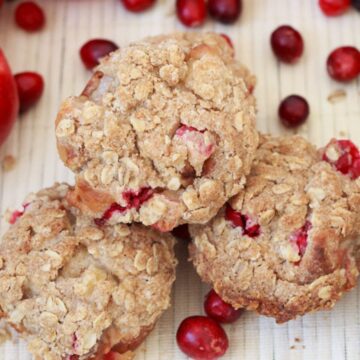 Three crumb-topped breakfast breads are placed on a light-colored surface surrounded by red apples, green apples, and scattered red cranberries. The muffins appear to have an oatmeal topping with visible pieces of cranberries inside.