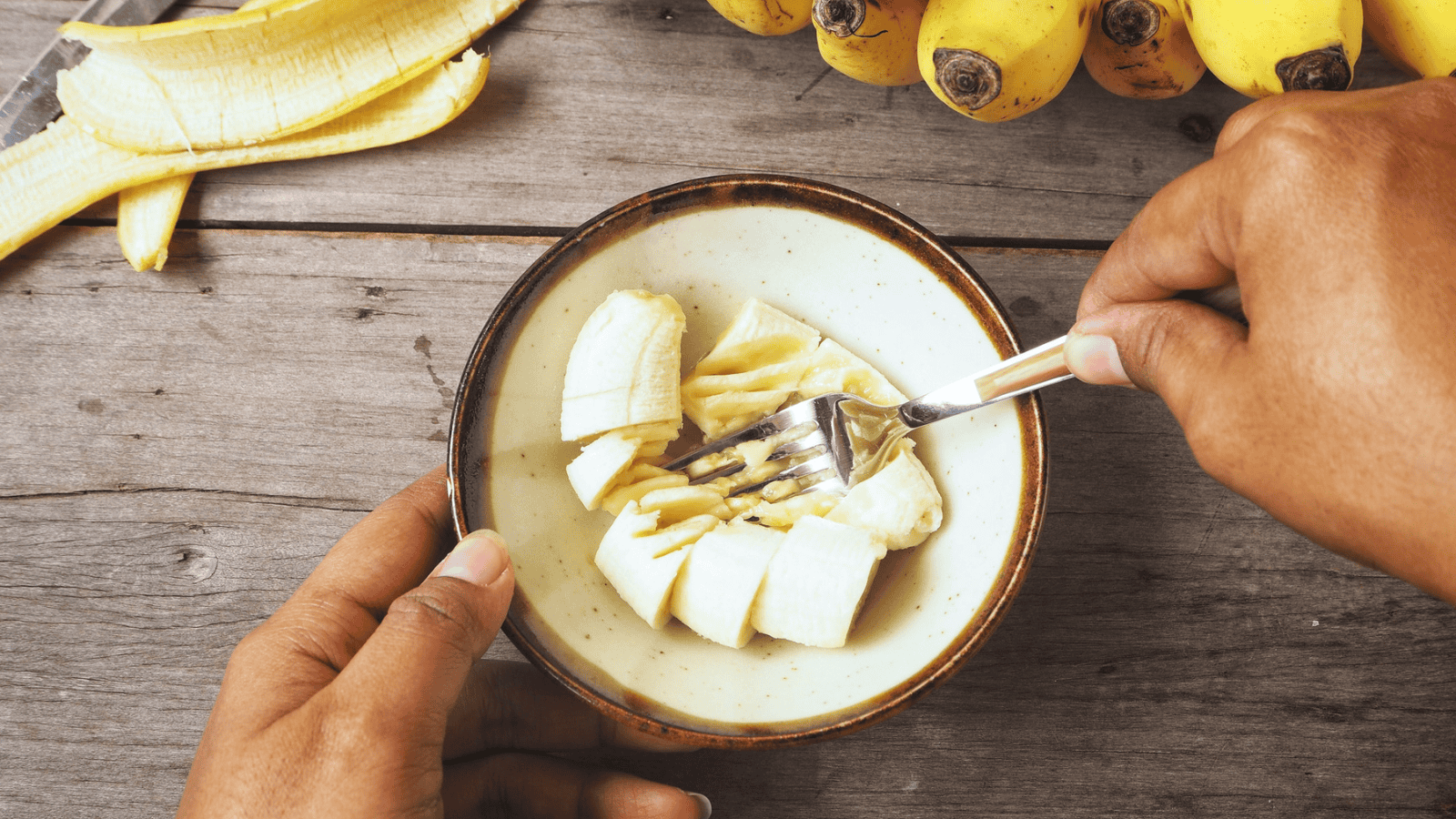 A person mashing banana slices in a bowl with a fork. The table holds additional bananas, both peeled and unpeeled, and a knife is visible in the background. The person is using both hands to hold the bowl and fork.