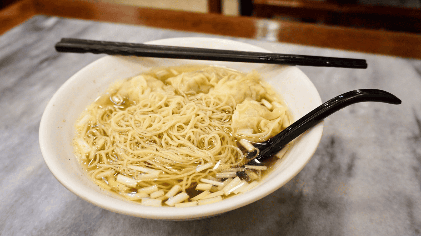 A bowl of noodle soup is topped with several dumplings and chopped green onions. A pair of black chopsticks and a black soup spoon rest on the edge of the white bowl. The bowl is placed on a table with a light-colored surface.