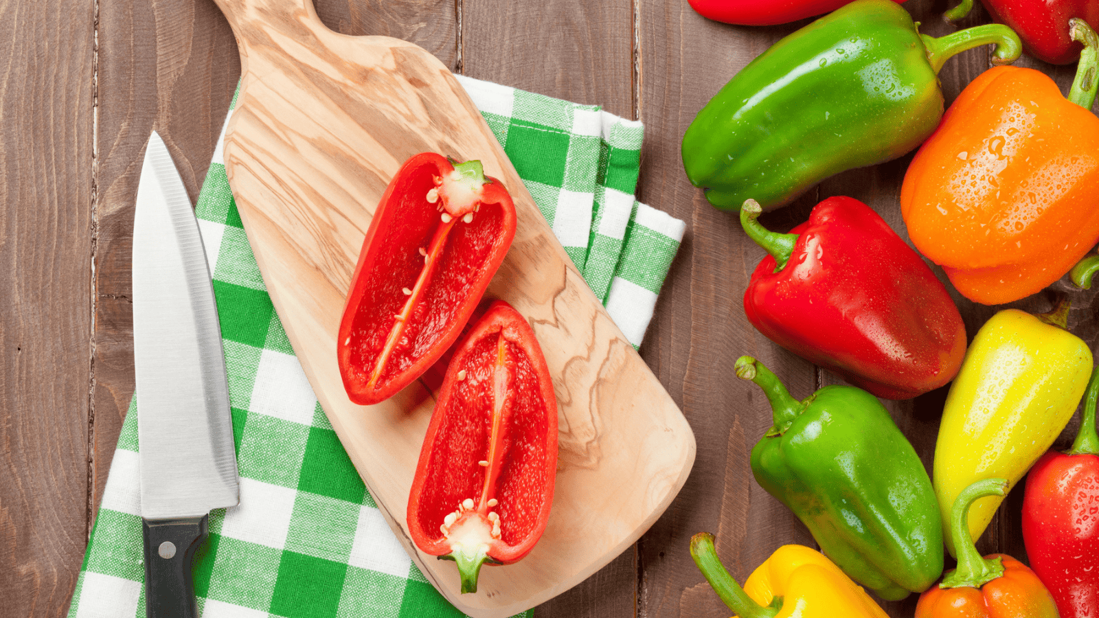 A wooden cutting board on a green and white checkered cloth holds a red bell pepper sliced in half next to a kitchen knife. To the right, there is a variety of whole bell peppers in red, green, yellow, and orange.