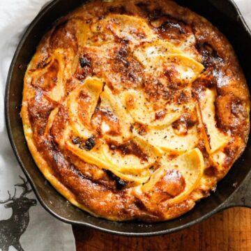 A freshly baked frittata in a black skillet sits on a wooden surface. The golden brown delight, with visible slices of apple or potato and raisins or dried fruit on top, hails from Grandpa's recipes. Beside the skillet, a white cloth with a black deer silhouette adds charm to the scene. It's begging for more!