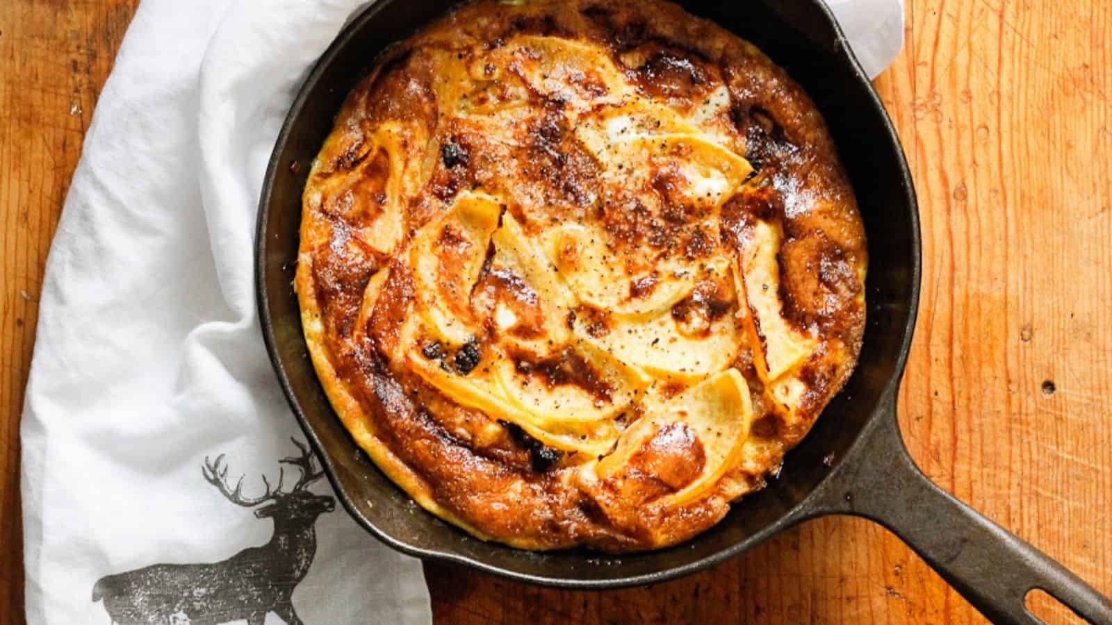 A freshly baked frittata in a black skillet sits on a wooden surface. The golden brown delight, with visible slices of apple or potato and raisins or dried fruit on top, hails from Grandpa's recipes. Beside the skillet, a white cloth with a black deer silhouette adds charm to the scene. It's begging for more!