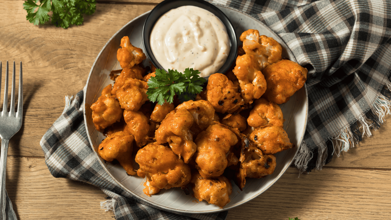 A plate of buffalo cauliflower wings garnished with parsley is served with a side dish of creamy dipping sauce. The plate is placed on top of a neatly folded checkered napkin on a wooden surface, with a silver fork positioned nearby.
