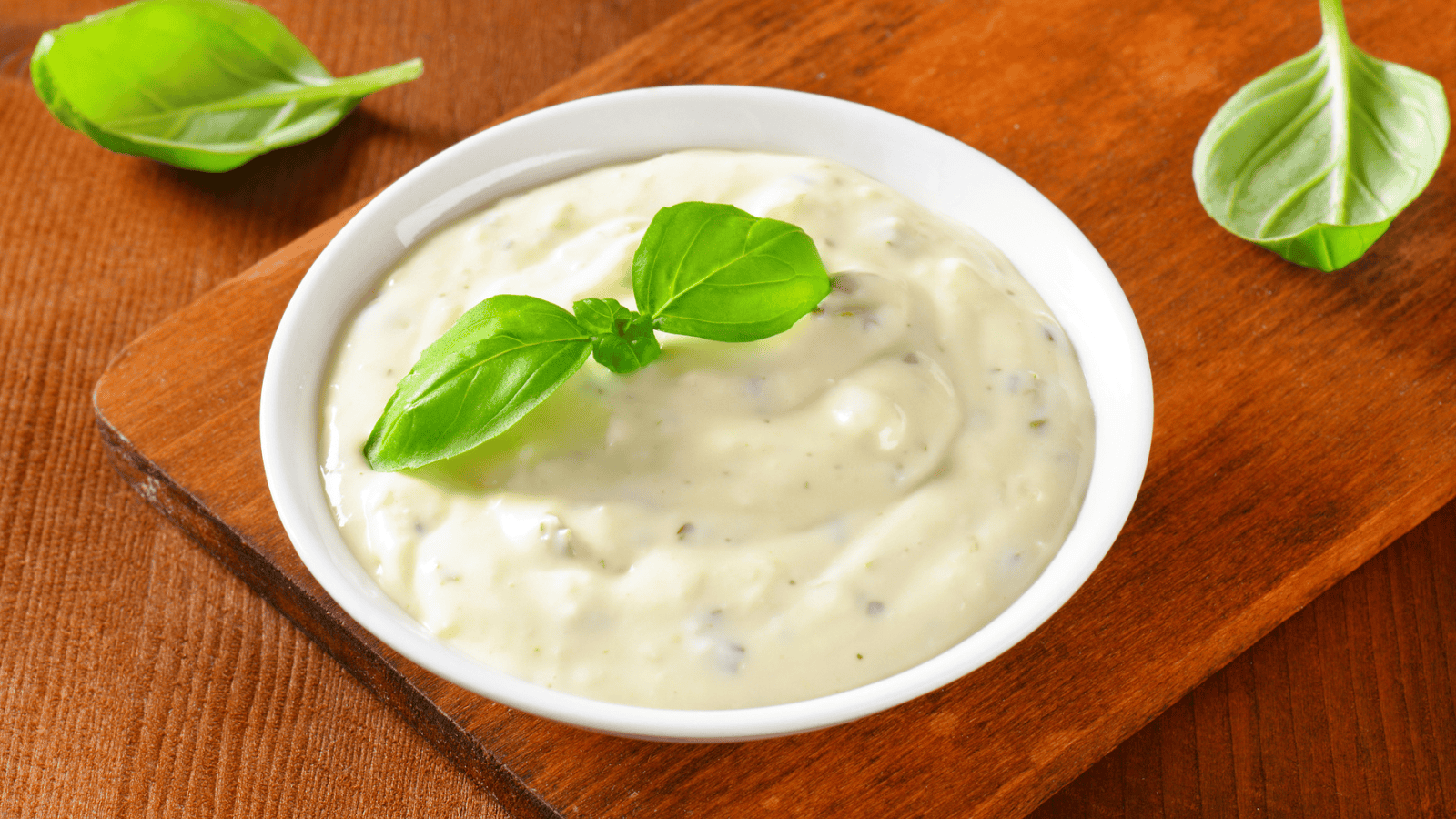 A bowl of creamy, white sauce garnished with a sprig of fresh basil is placed on a wooden surface. Three loose basil leaves adorn the background. The sauce has a smooth texture and is served in a small white bowl.