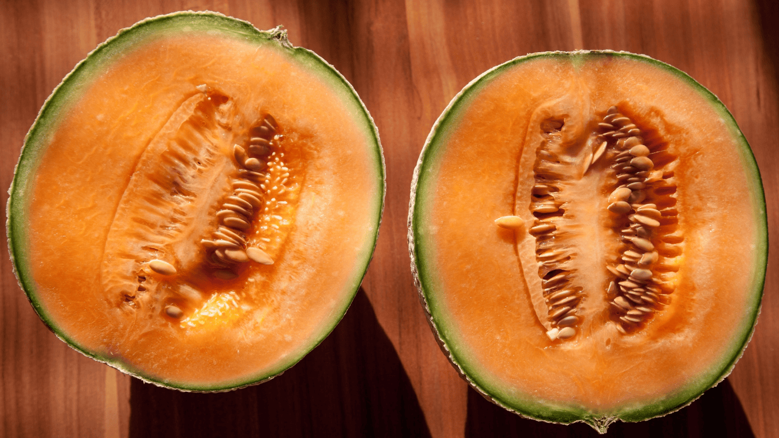 A close-up of a ripe cantaloupe cut in half, showing the orange flesh and seeded center. The melon's green rind encircles the orange interior, which has a slightly rough texture. The image is taken on a wooden surface.