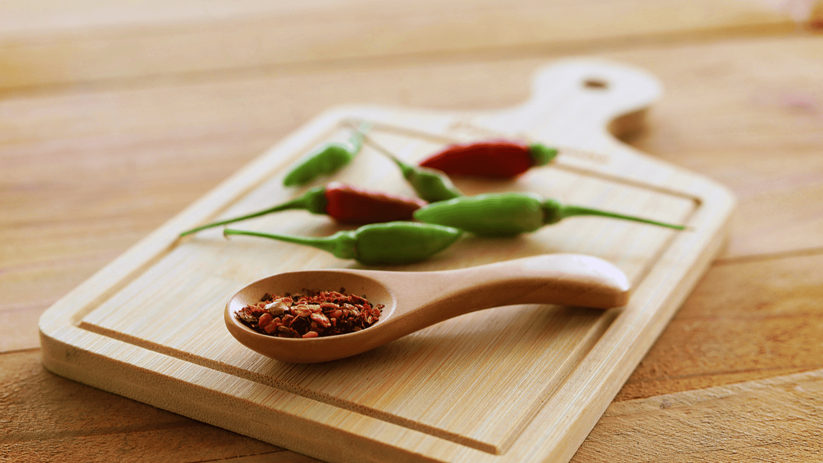 A wooden cutting board with a handle features a wooden spoon filled with crushed red pepper flakes. Several fresh green and red chili peppers are also on the cutting board. The background is a wooden surface.