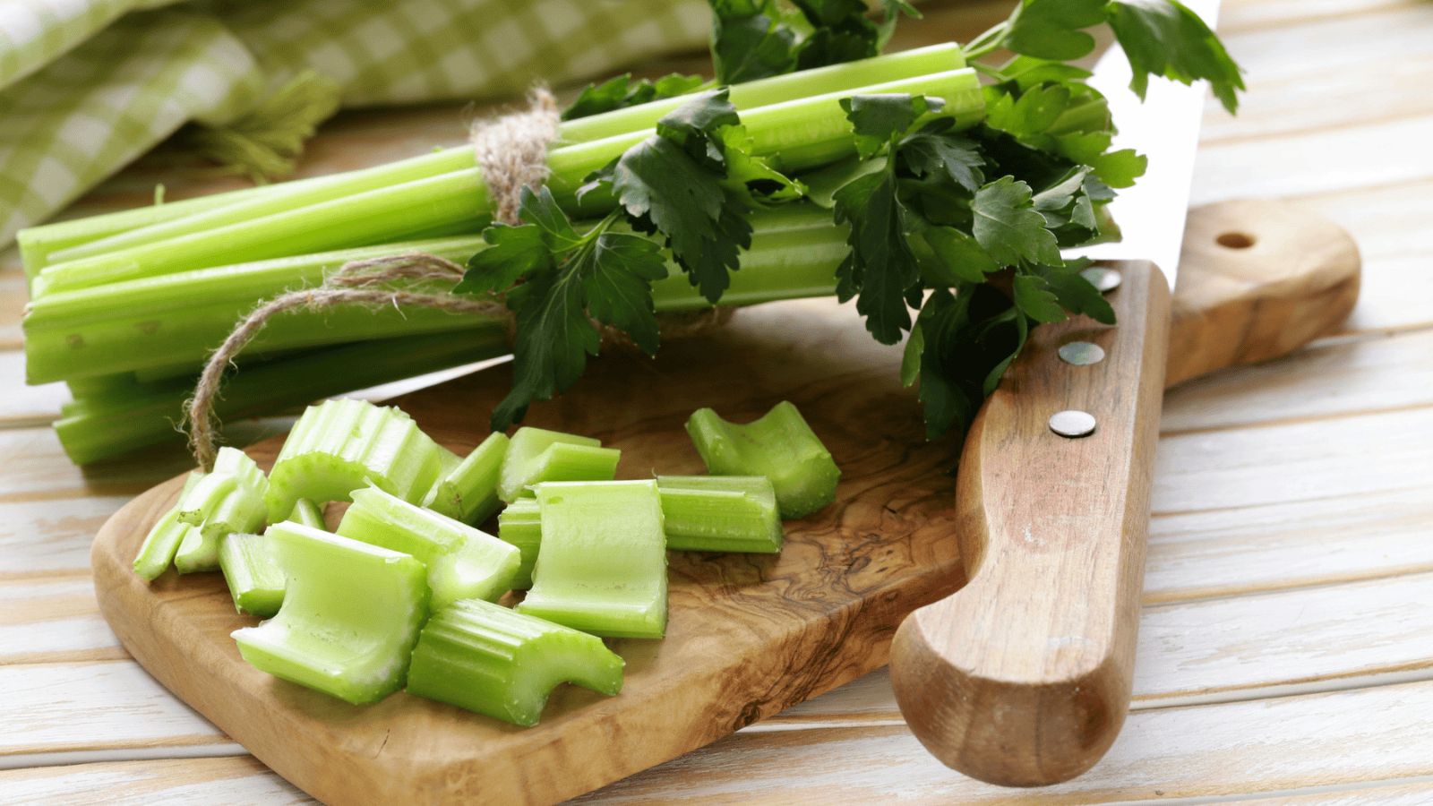 A bundle of celery with leaves is tied together with twine and placed next to a large knife on a wooden cutting board. Some chopped celery pieces are also on the cutting board. A green and white checkered cloth is in the background.