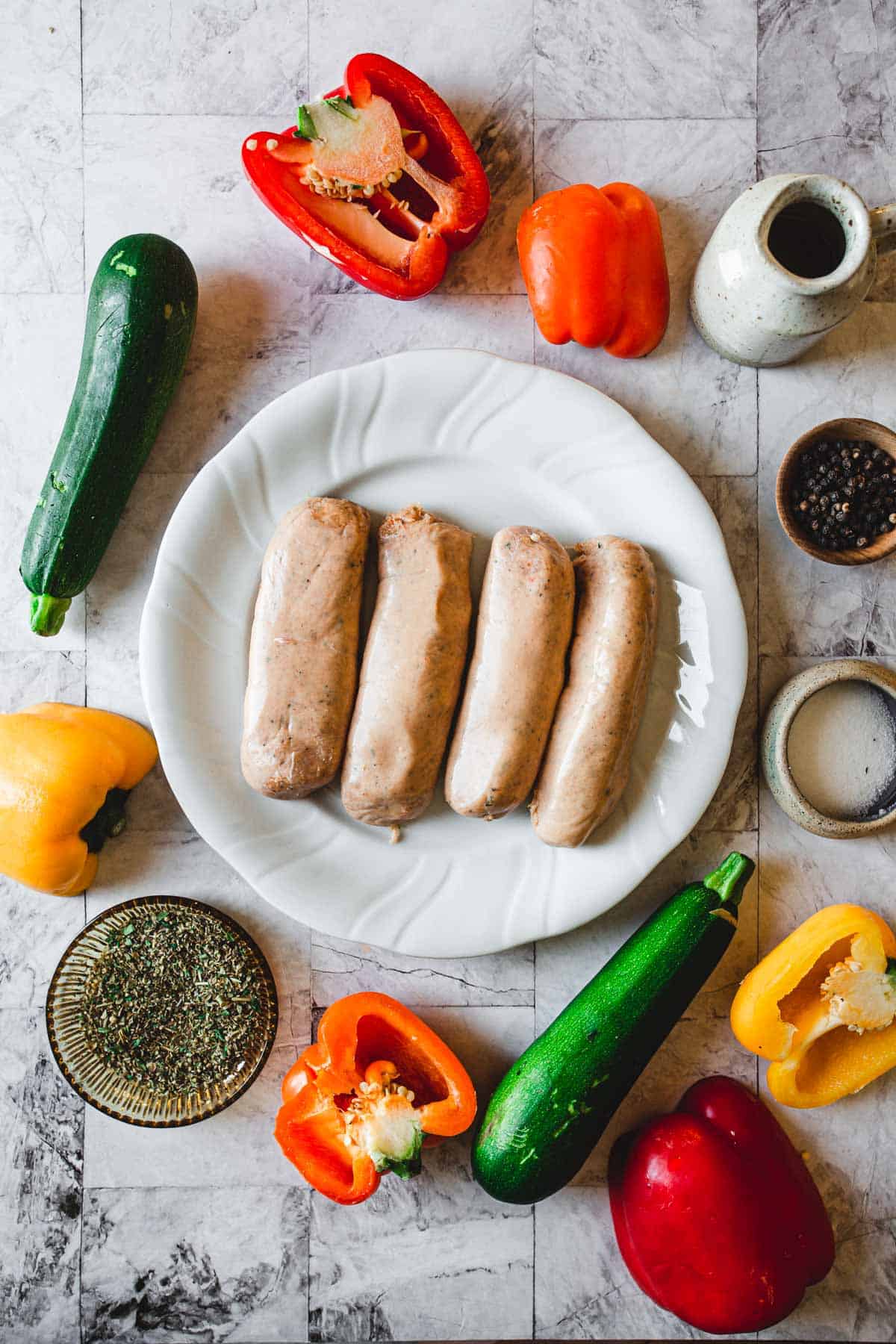 A plate with four sausages is centered on a marble surface, surrounded by cut red, orange, and yellow bell peppers, two zucchinis, a bowl of spices, a jar of peppercorns, a small bowl of salt.