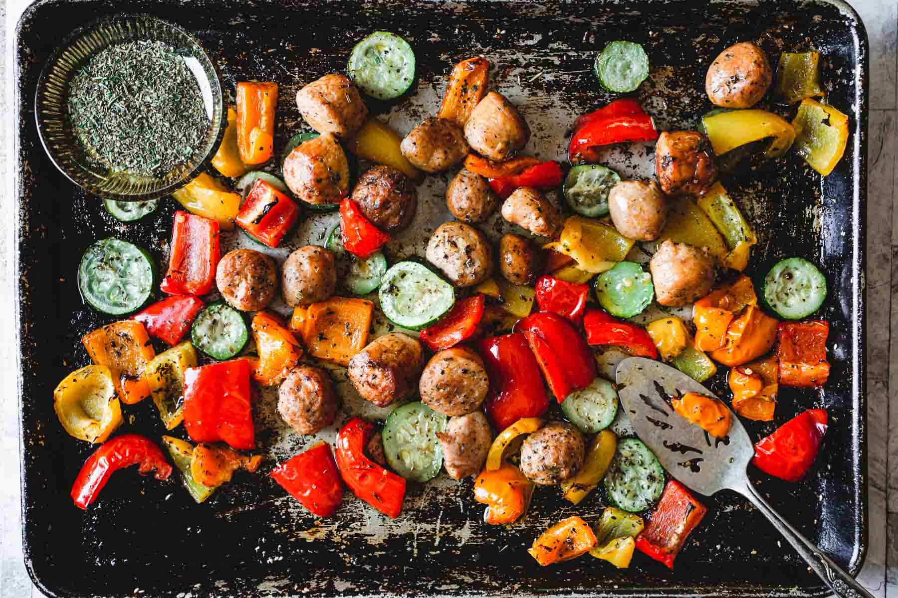 A well-used baking tray loaded with sausage and veggies—red and yellow bell peppers, sliced zucchini, and cooked sausage pieces. A metal serving utensil rests beside a small glass bowl of seasoning in the top left corner. Perfect for an easy weeknight dinner.