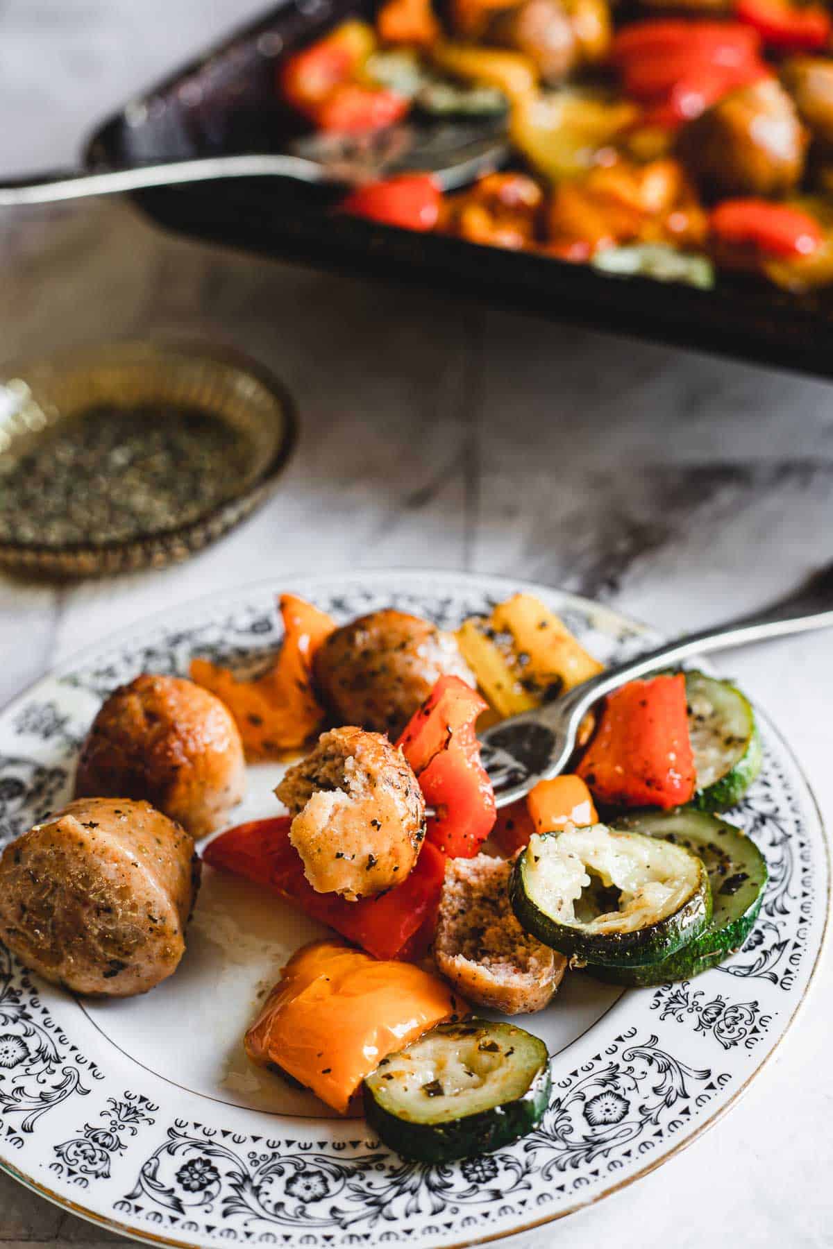 A white plate with black decorative patterns holds a serving of roasted veggies and sausage pieces. Vegetables include red and yellow bell peppers and zucchini slices. A fork is placed on the plate, and a baking tray with more food is visible in the background – perfect for an easy weeknight dinner.