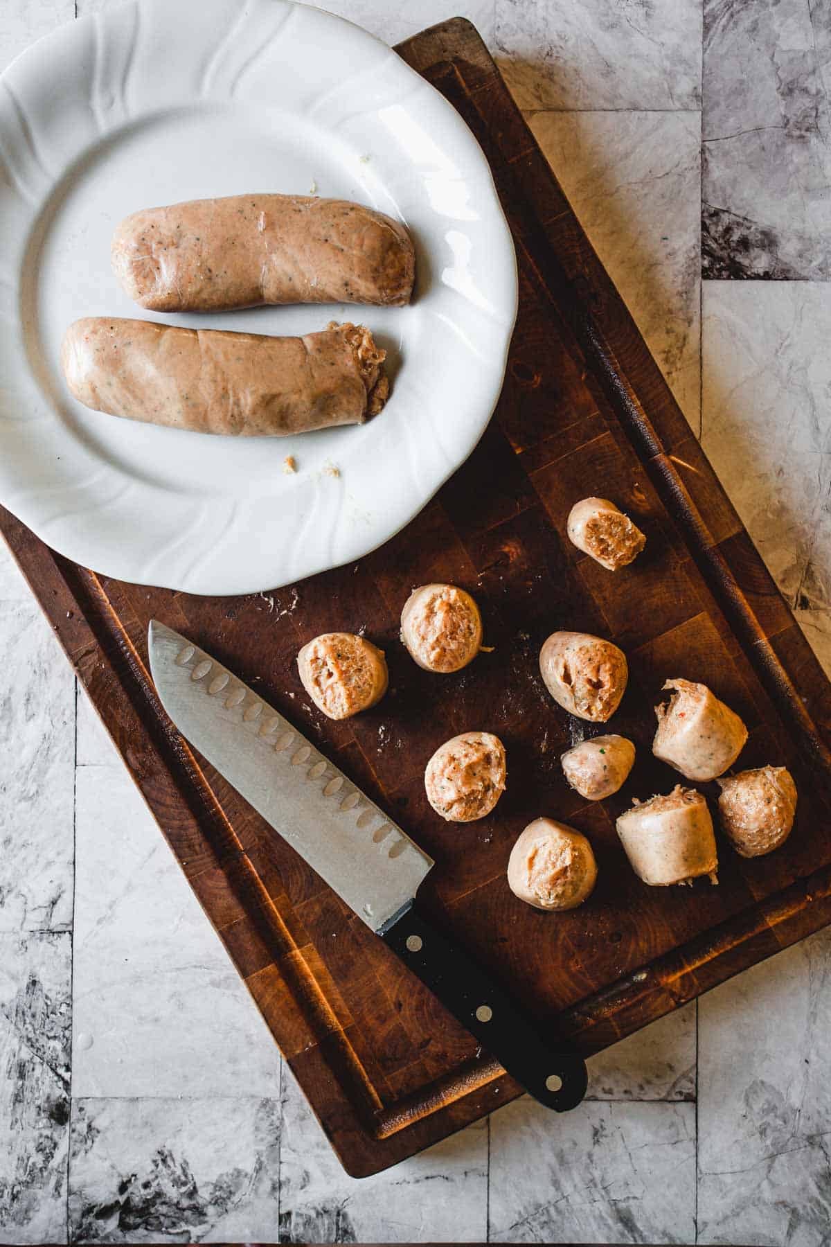 A wooden cutting board featuring a sharp knife, sliced sausages, and two whole sausages arranged on a white plate beside it is set on a marble countertop.