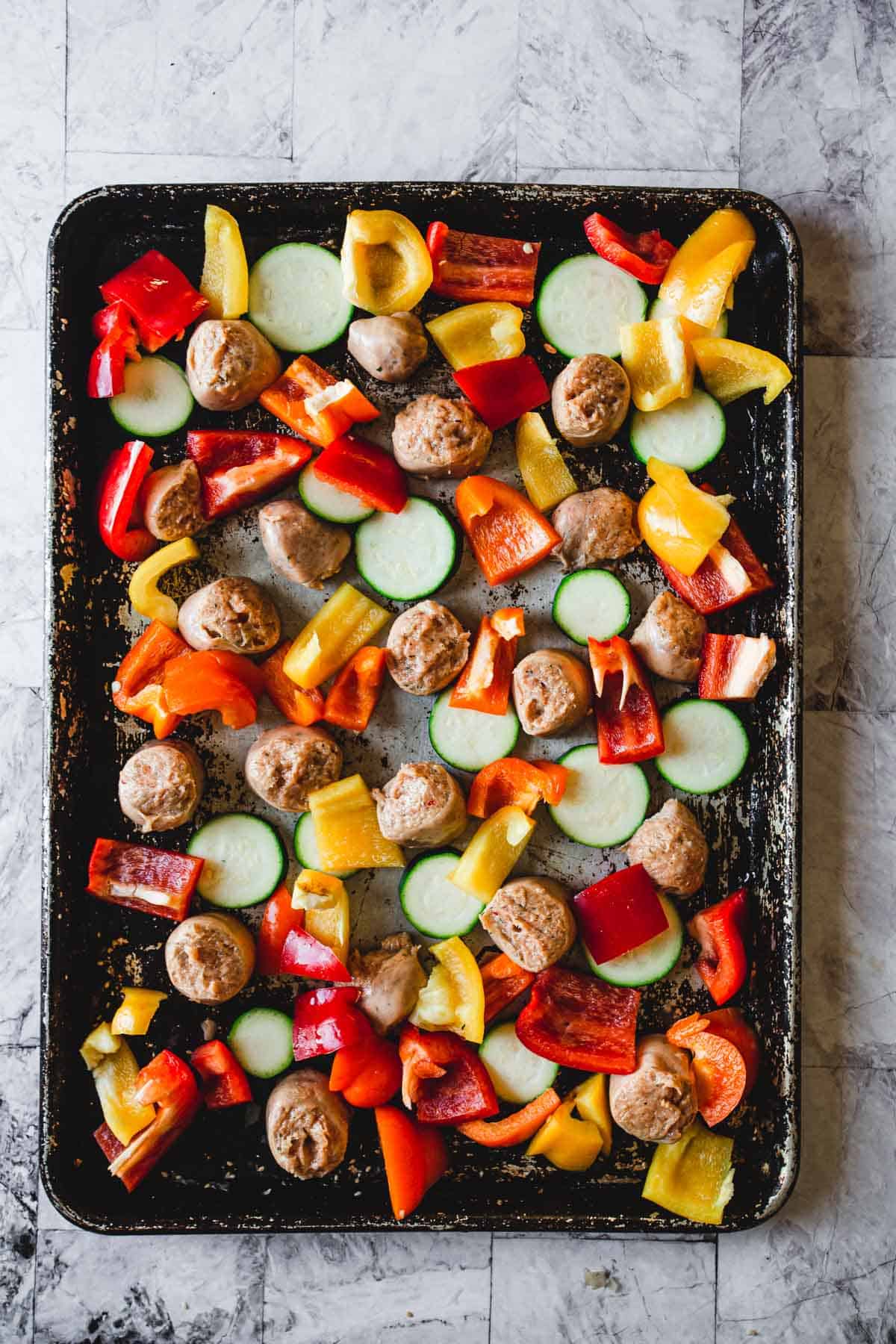 A baking sheet filled with sliced zucchini, diced red and yellow bell peppers, and small meatballs is prepped for roasting. All arranged on a dark, seasoned baking tray.