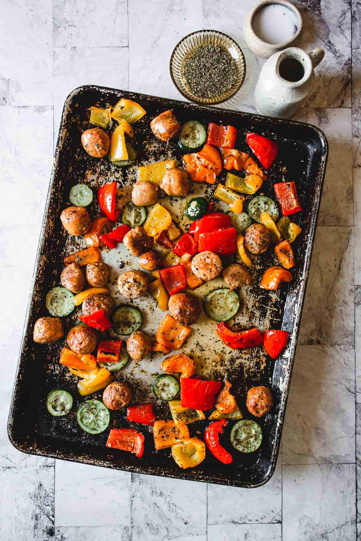 A baking tray filled with roasted vegetables and sausage pieces makes for an easy weeknight dinner. The vegetables include sliced zucchini, red bell peppers, and yellow bell peppers. A small bowl of spices and a white sauce pitcher are placed beside the tray on a marble surface.