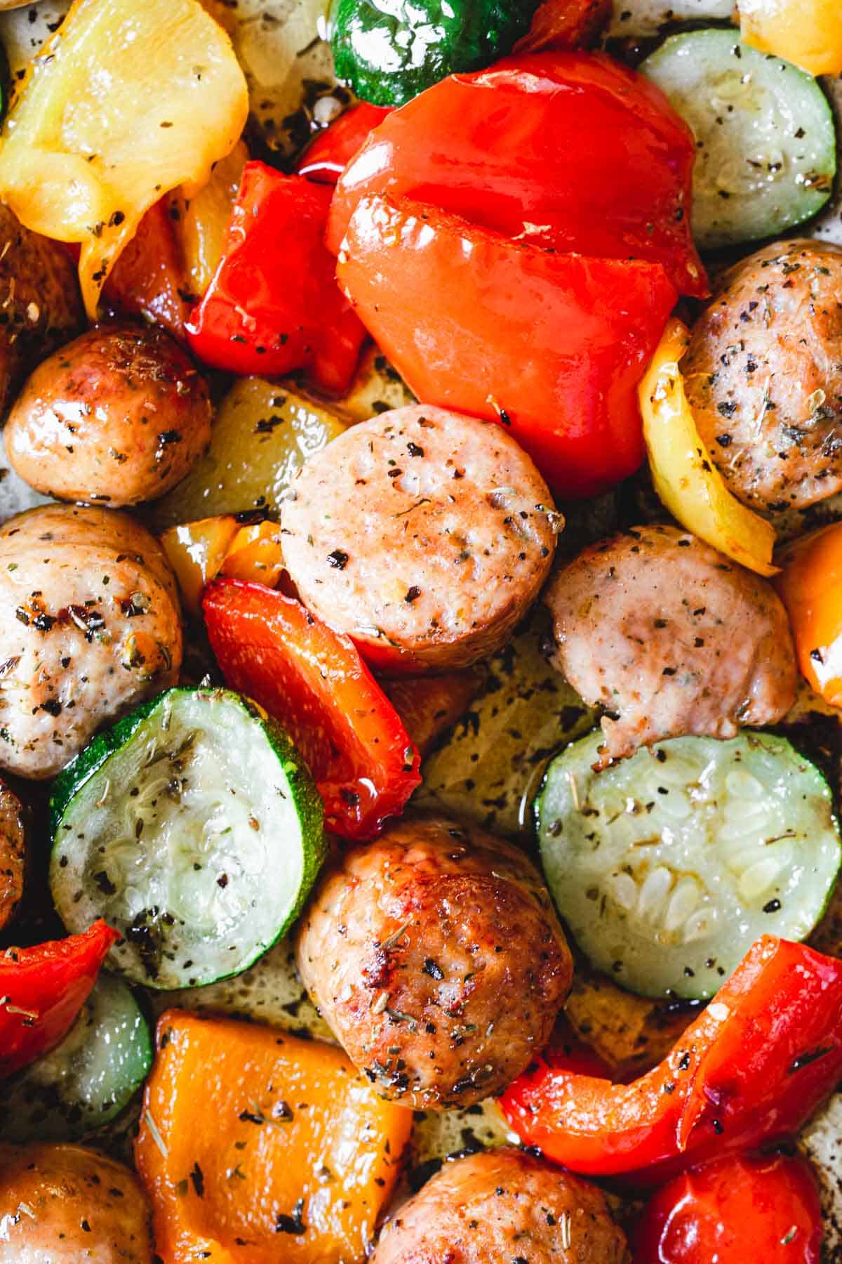 A close-up of a roasted dish featuring pieces of sausage, zucchini, and red and yellow bell peppers, all seasoned with herbs. This delicious veggie dinner shows the ingredients are well-cooked with a slight char on the edges.