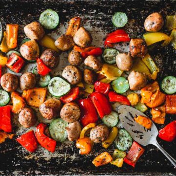 A baking sheet with chicken sausage, roasted vegetables, and a medley of red, orange, and yellow bell peppers alongside sliced zucchini. A metal slotted spatula is placed on the right side of the sheet pan.