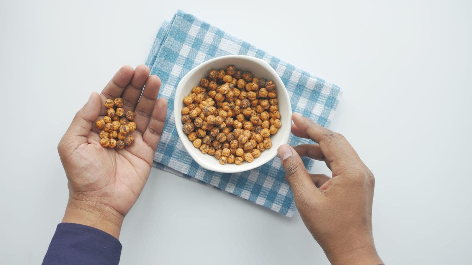 A person's hands holding roasted chickpeas over a blue and white checkered cloth, with one hand holding a white bowl filled with more chickpeas. The background is a plain white surface. Discover Veggie-Powered Proteins!