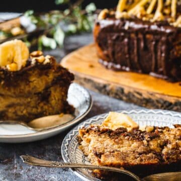 Two plates with slices of banana bread, perfect for those who enjoy breakfast breads, are on a table. Each slice has a piece of banana on top. Behind the plates is a wooden board with additional slices of banana bread. Greenery is used as decoration around the table setting.