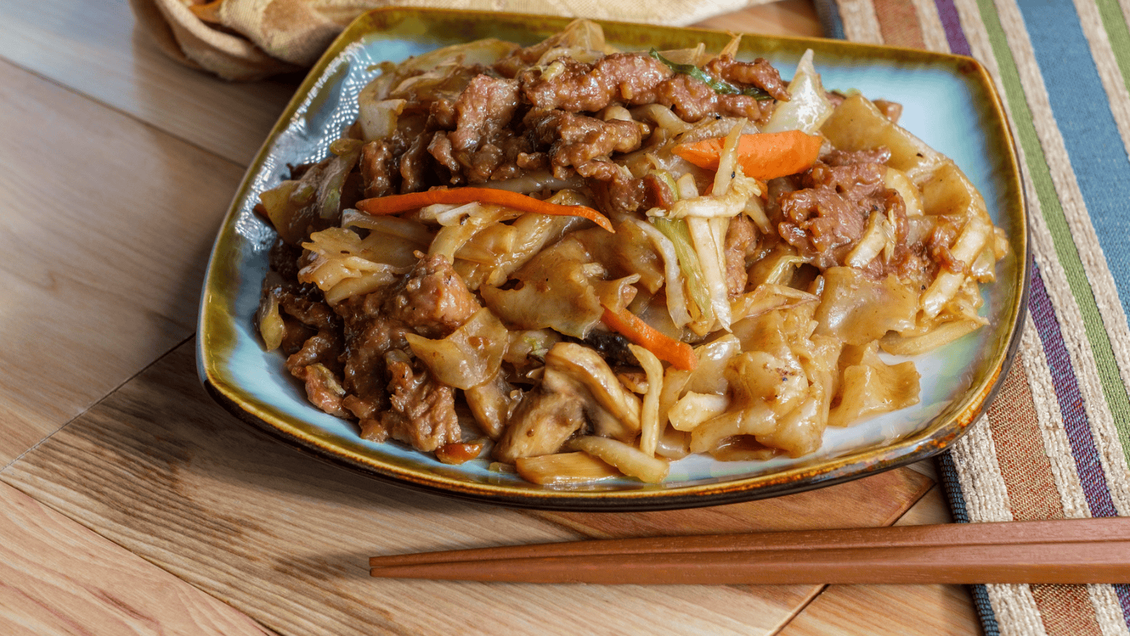 A square plate filled with stir-fried noodles, sliced beef, shredded cabbage, and carrot strips is placed on a wooden table. Brown wooden chopsticks rest beside the plate, and a colorful striped fabric and a beige cloth are in the background.