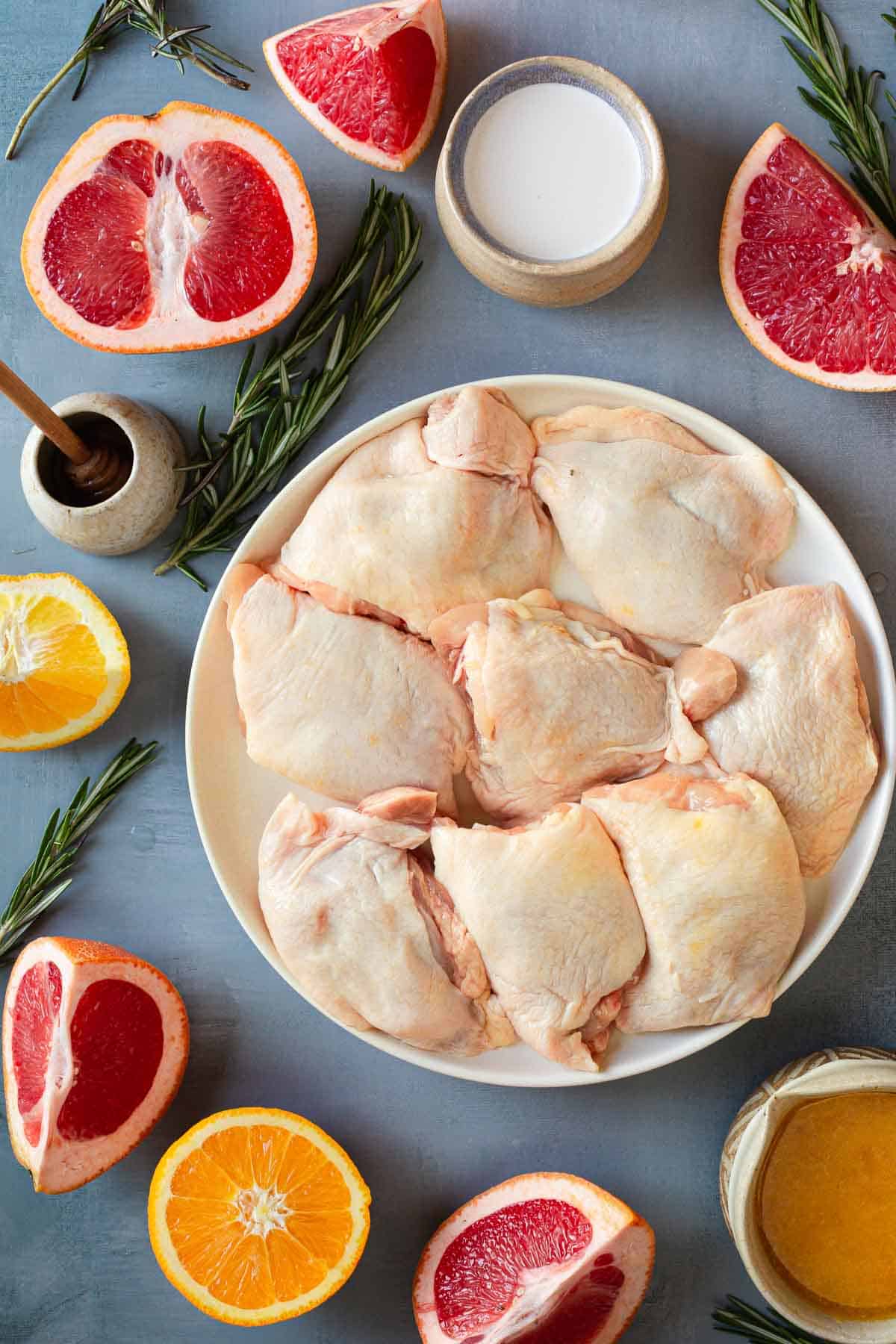 A plate of raw chicken thighs is surrounded by halved grapefruits, halved oranges, rosemary sprigs, a small dish of honey with a honey dipper, a bowl of salt, and a bowl of sauce. The food items are arranged on a light blue surface, ready to be transformed into creamy citrus chicken.