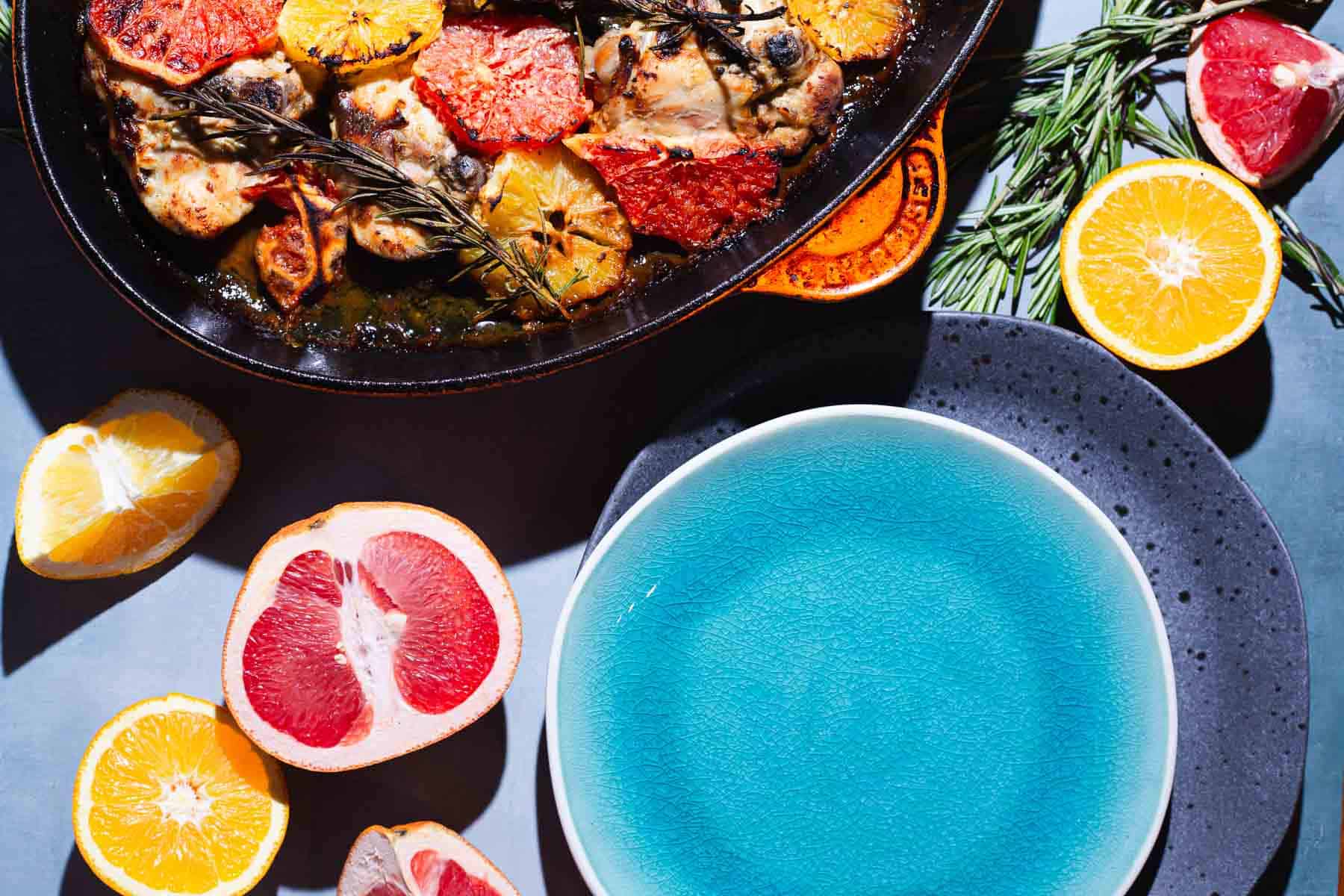 A ceramic dish with cooked creamy citrus chicken pieces garnished with citrus slices and rosemary sits next to a blue plate. Surrounding the dish and plate are halved citrus fruits, including grapefruit, orange, lemon, and lime, arranged on a gray surface.