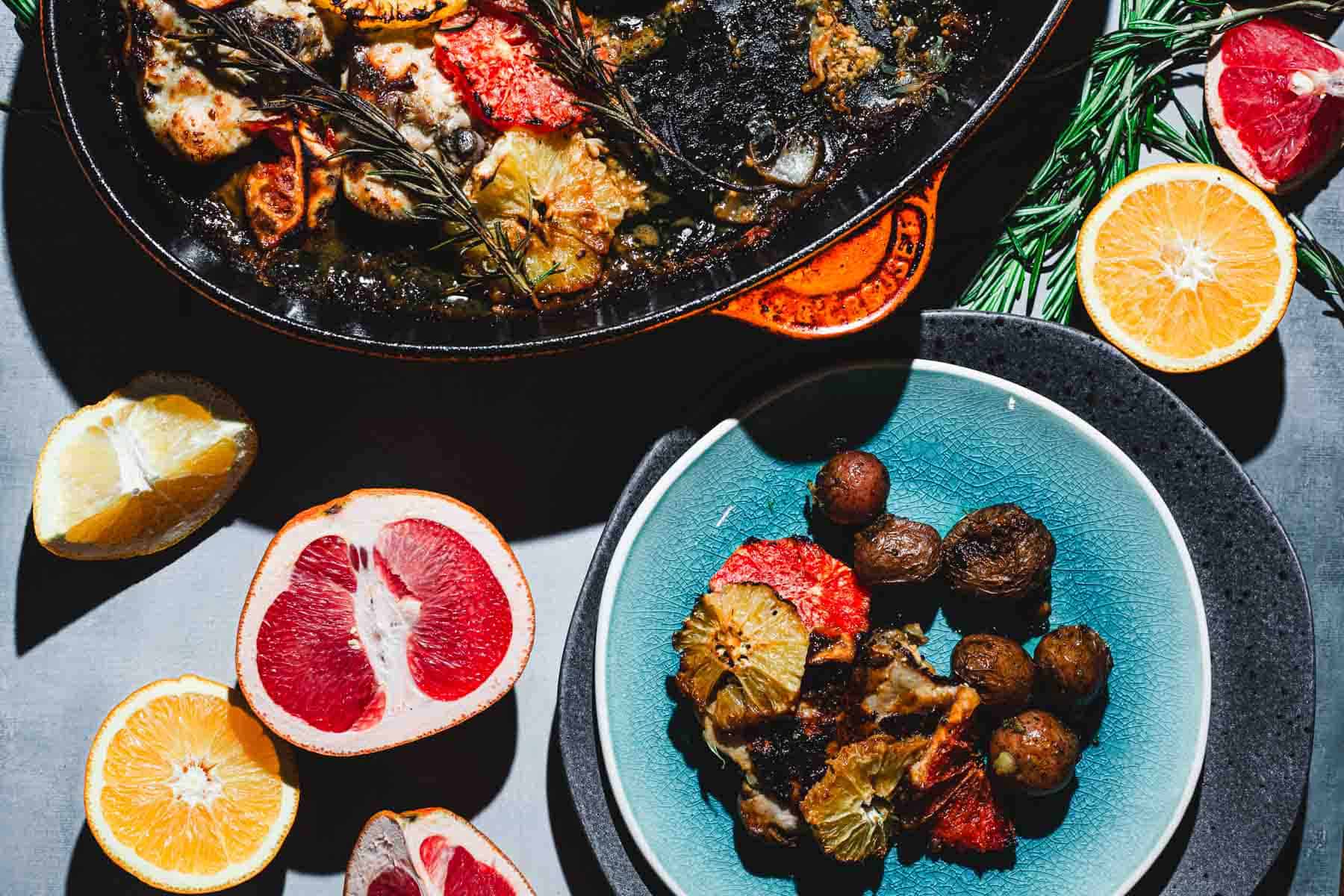 An overhead view of a meal featuring a dish of cooked vegetables and small potatoes on a blue plate and another dish with creamy citrus chicken, garnished with rosemary. Surrounding the dishes are sliced citrus fruits, including grapefruit and lemon.