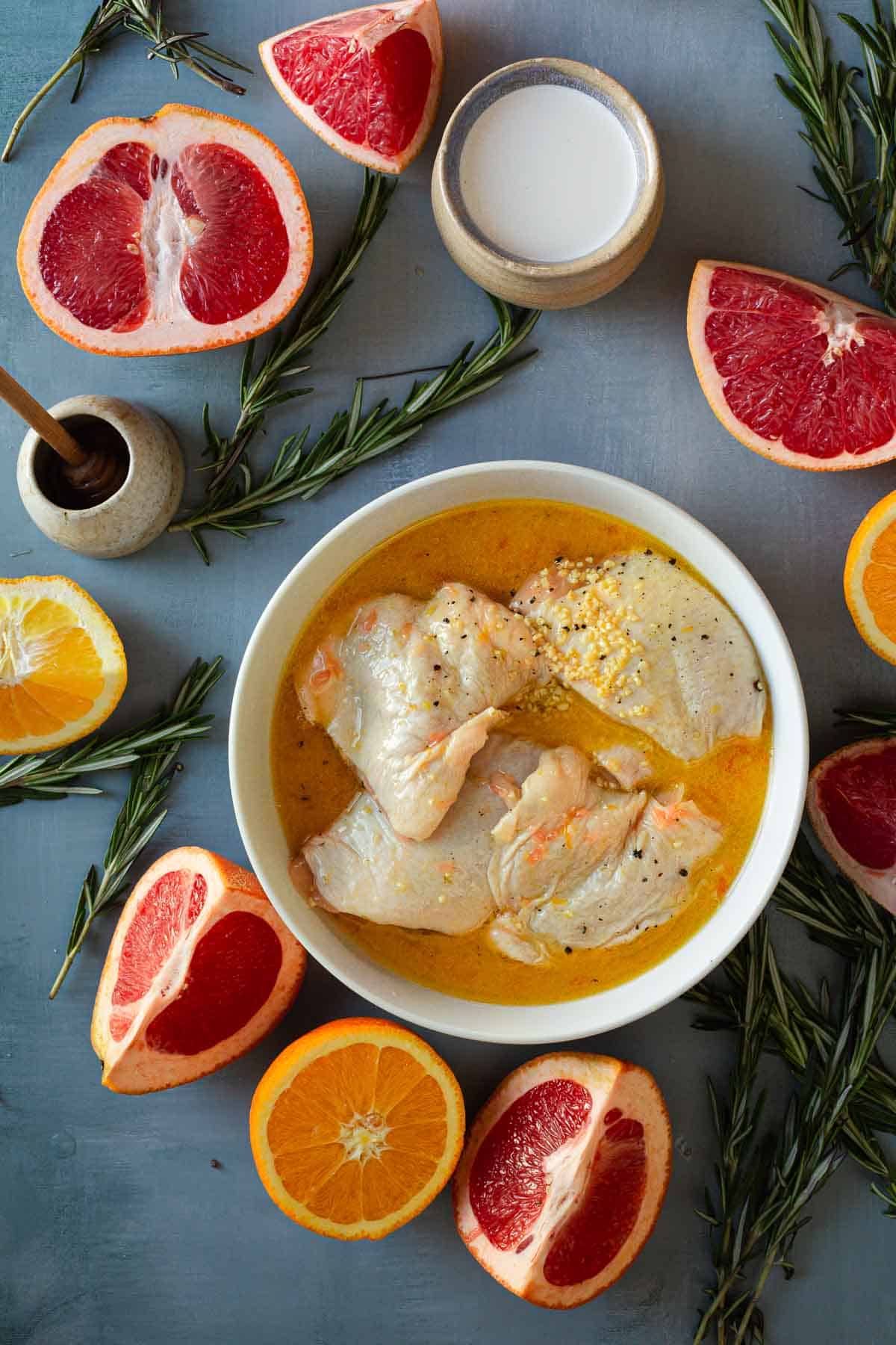 A bowl of raw chicken pieces marinating in a yellow mixture, surrounded by halved grapefruits, oranges, sprigs of rosemary, a small container of white liquid, and a honey dipper in a small pot. The ingredients for this delicious Citrus Chicken are arranged on a blue surface.