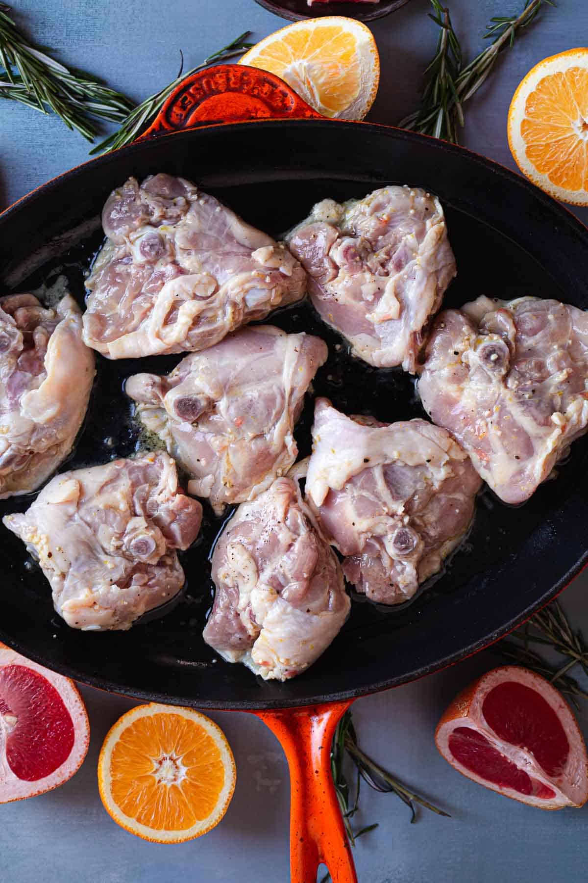 A frying pan with raw chicken pieces arranged inside it. Surrounding the pan are various citrus halves, including orange and grapefruit, along with sprigs of rosemary. This setup promises a perfect creamy citrus chicken dish on a flat surface.