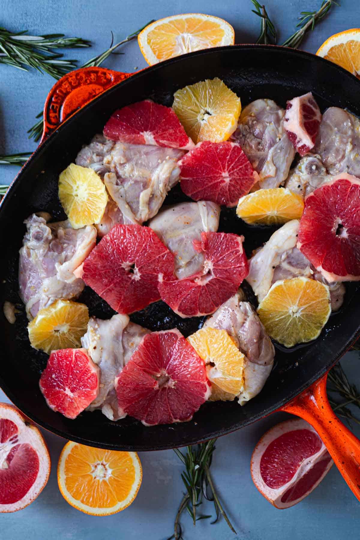 A black skillet contains raw chicken pieces topped with slices of red, yellow, and orange citrus fruits. The skillet sits on a blue surface, surrounded by additional citrus slices and sprigs of rosemary. An orange cooking utensil is visible on the left side, ready to create Perfect Creamy Chicken.