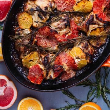 A baked dish in an oval-shaped orange pan, featuring creamy citrus chicken pieces garnished with slices of grapefruit, orange, and lemon, along with sprigs of rosemary. Halved citrus fruits and rosemary sprigs are scattered around the pan on a blue surface.