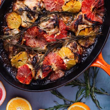 A close-up of a baked dish featuring Perfect Creamy Chicken thighs garnished with slices of orange and grapefruit, rosemary sprigs, and a dark glaze. The dish is in an orange baking tray, surrounded by rosemary sprigs and citrus slices on a gray surface.