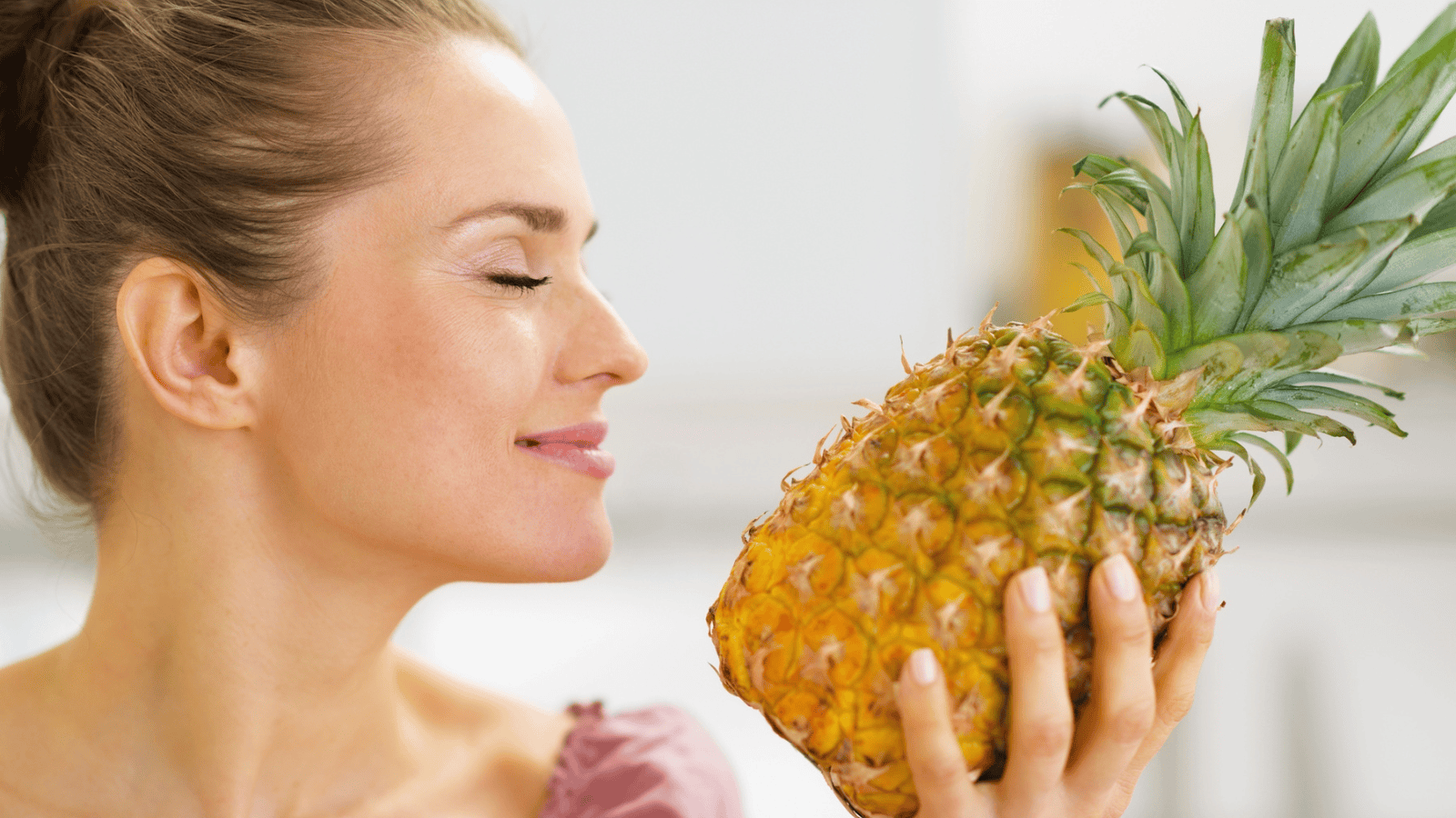 A woman with her hair tied back smiles with her eyes closed, holding a pineapple close to her face. The background is blurred, suggesting an indoor setting.