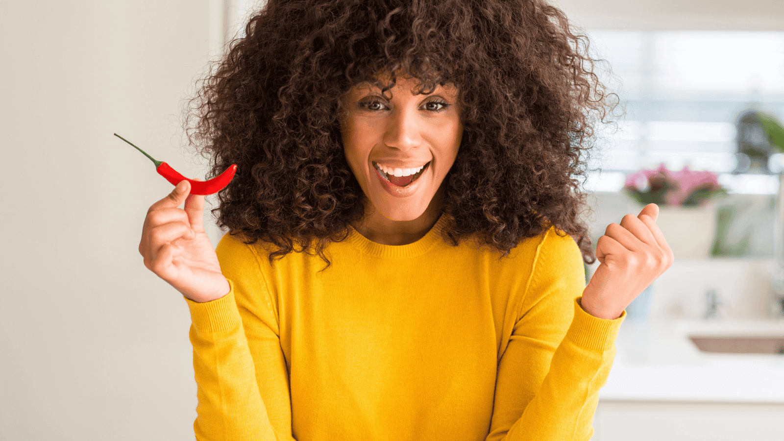 A person with curly hair wearing a yellow sweater is holding a red chili pepper in one hand and making a fist with the other. They are standing in a brightly lit kitchen and smiling at the camera.