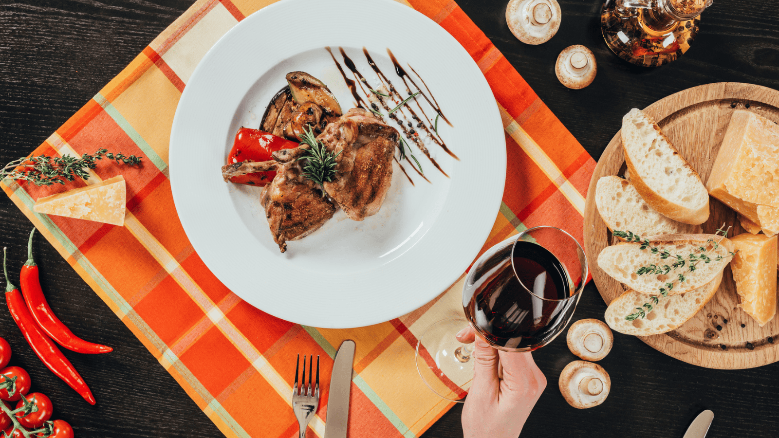 A white plate with grilled meat and vegetables drizzled with sauce sits on an orange plaid tablecloth. A hand holds a glass of red wine. Bread, cheese, and a bottle of oil are placed nearby.