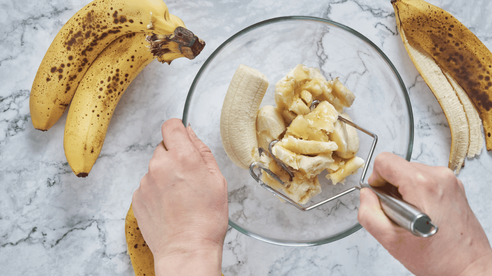 Two hands are mashing a peeled banana in a glass bowl using a fork. Two whole ripe bananas with brown spots and one partially peeled banana are on a marble surface around the bowl.