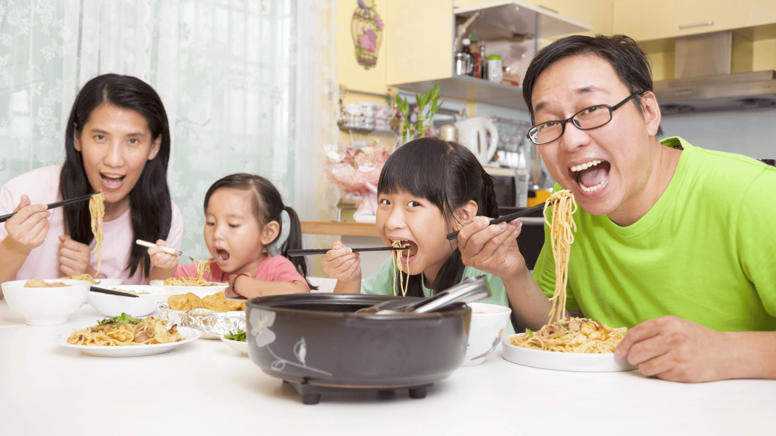 A family of four sits at a kitchen table, eating a meal together. The adults and children are using chopsticks to eat noodles and other dishes. The table is set with various plates of food and a black pot in the center. The background shows kitchen decor.