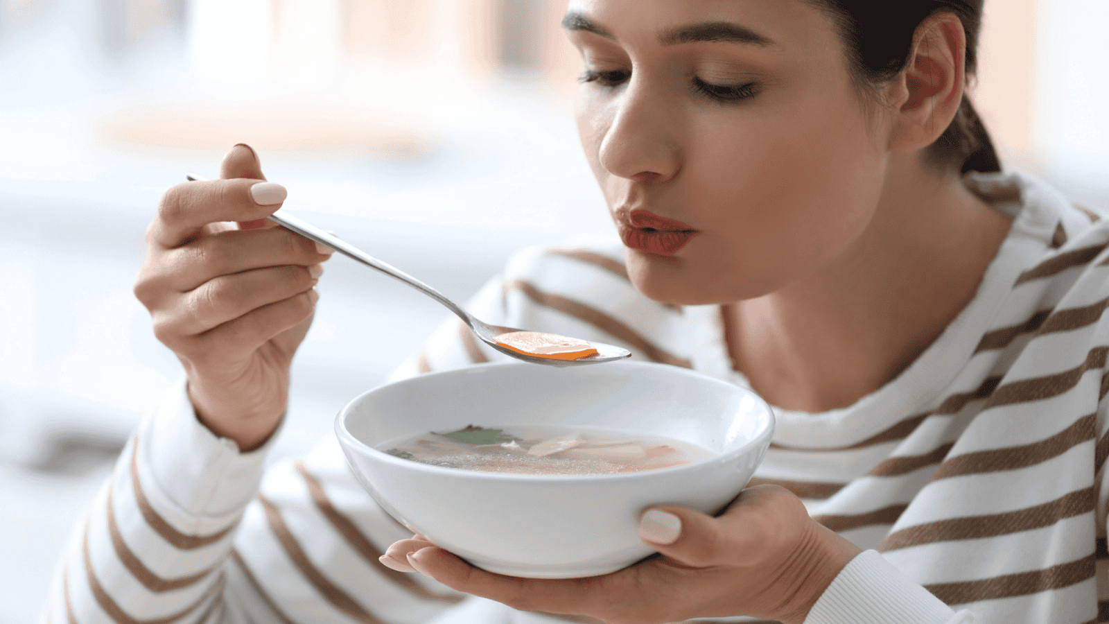A person with dark hair pulled back is blowing on a spoonful of soup held above a white bowl. The individual is wearing a white and tan striped shirt. The background is blurred, suggesting an indoor setting.