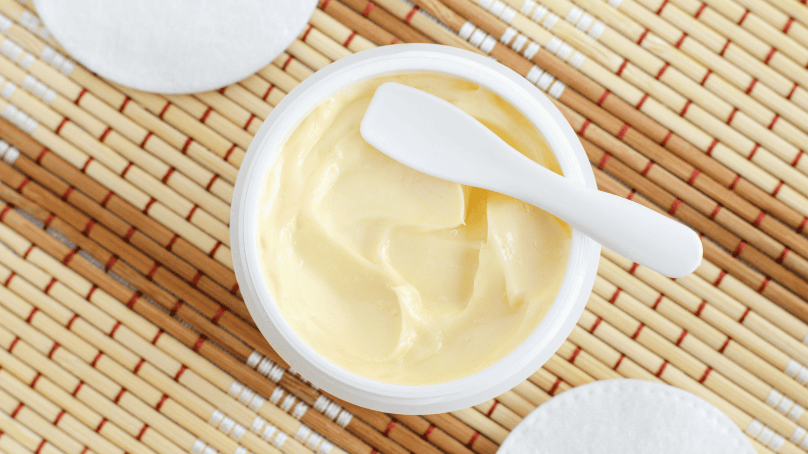 A close-up view of an open jar filled with a creamy yellow substance, with a white plastic spatula resting on top. The jar is placed on a woven bamboo mat, surrounded by round white pads.