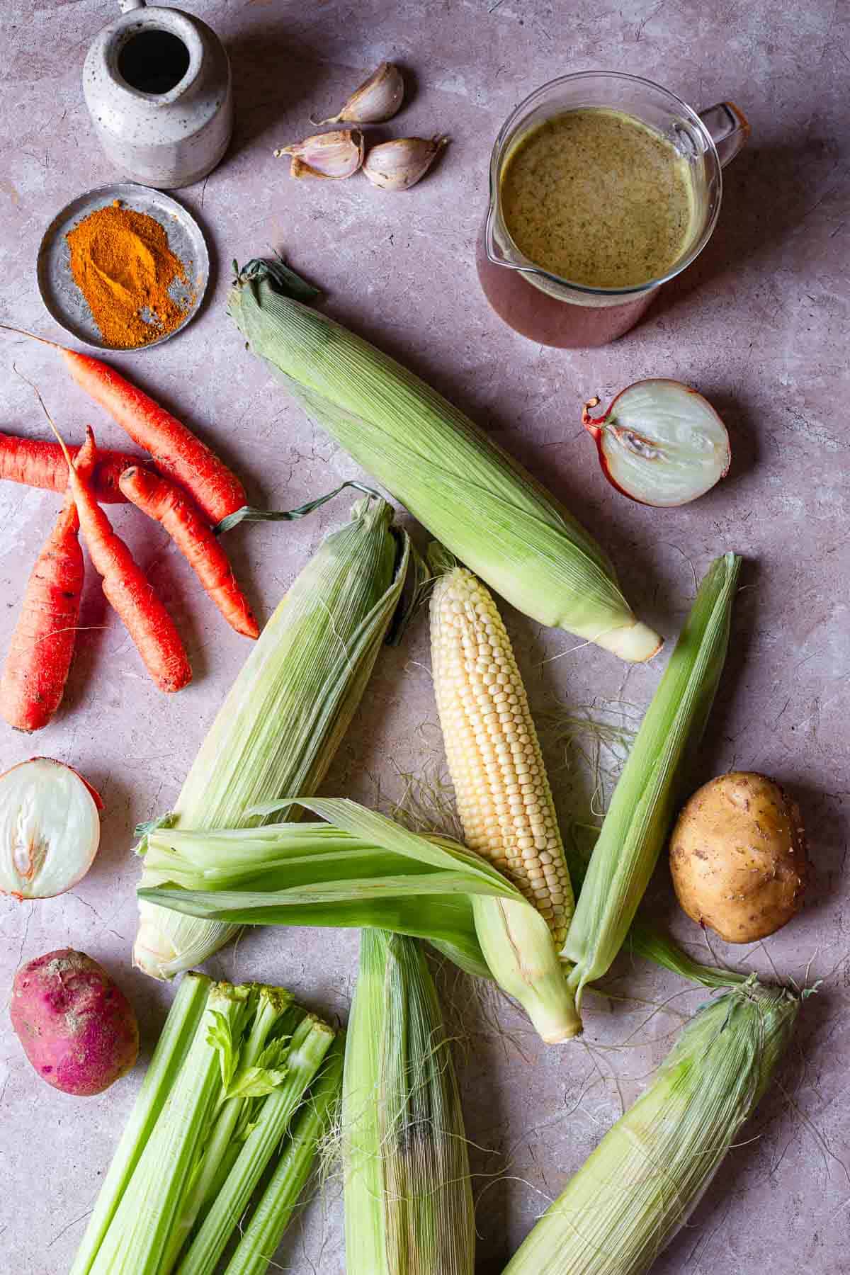 A variety of fresh vegetables and spices on a textured surface, including fresh corn on the cob, carrots, onions, garlic, and potatoes, accompanied by a small jug of sauce and ground turmeric.