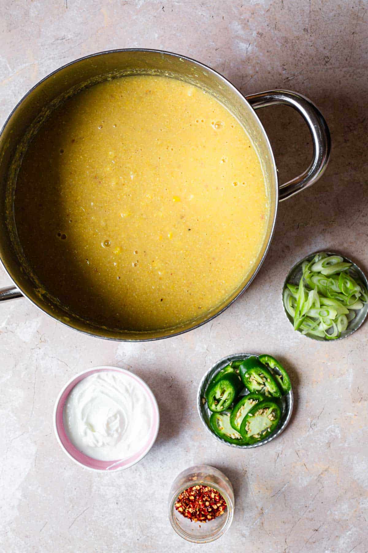 Top-down view of a pot of blended yellow corn soup, accompanied by small bowls of sliced jalapeños, sour cream, sliced green onions, and crushed red pepper on a gray countertop.